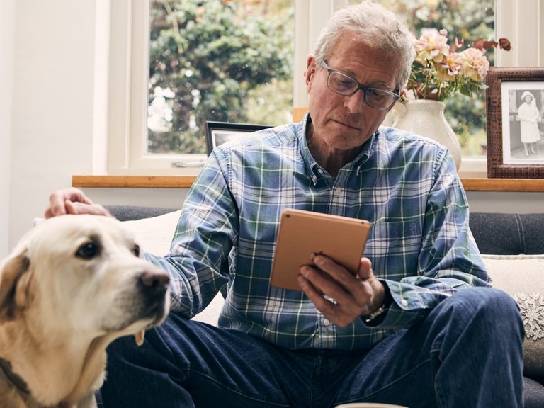 An older man looking at a tablet in his living room, with his dog by his side