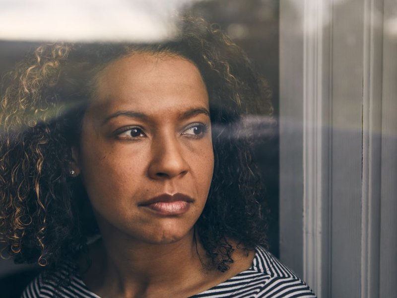 A woman with curly brown hair is staring out of a window with a thoughtful expression on her face