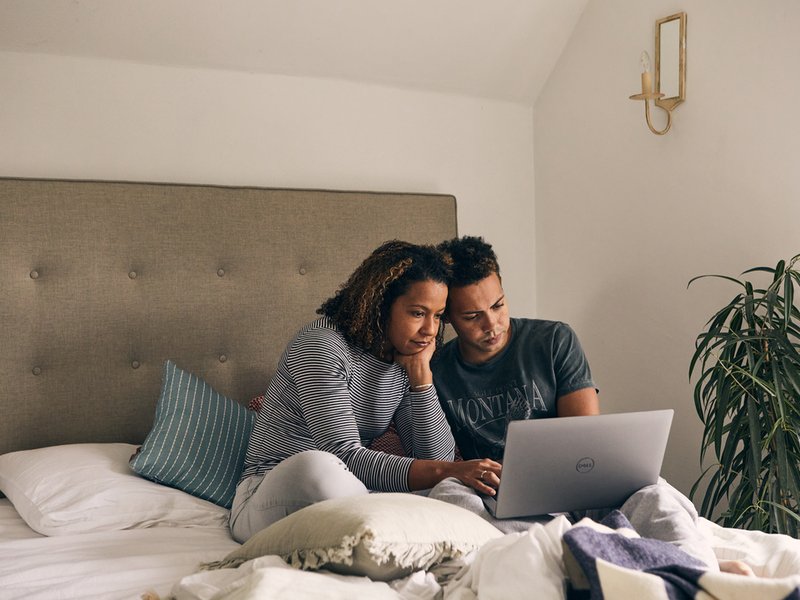 A black woman sits on a double bed next to a younger black male whilst they both look thoughtfully at a laptop. The son leans his head against his mother's.