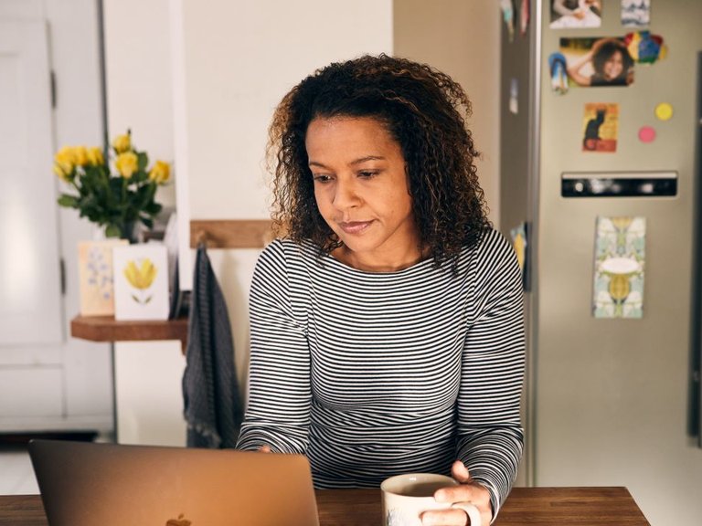 Women sits at kitchen table clutching a mug in one hand. She's looking at a laptop.