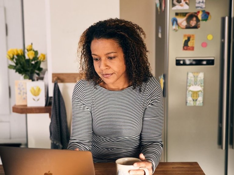 A woman is sat at the kitchen table looking at a laptop in front of her, holding a mug