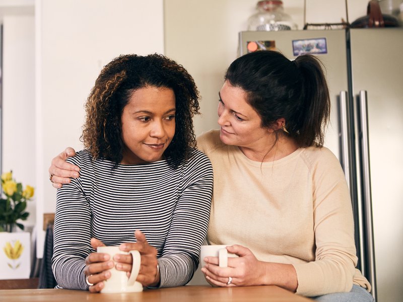 Two women sit in a kitchen with mugs. One woman has her arm around the other in a comforting gesture.