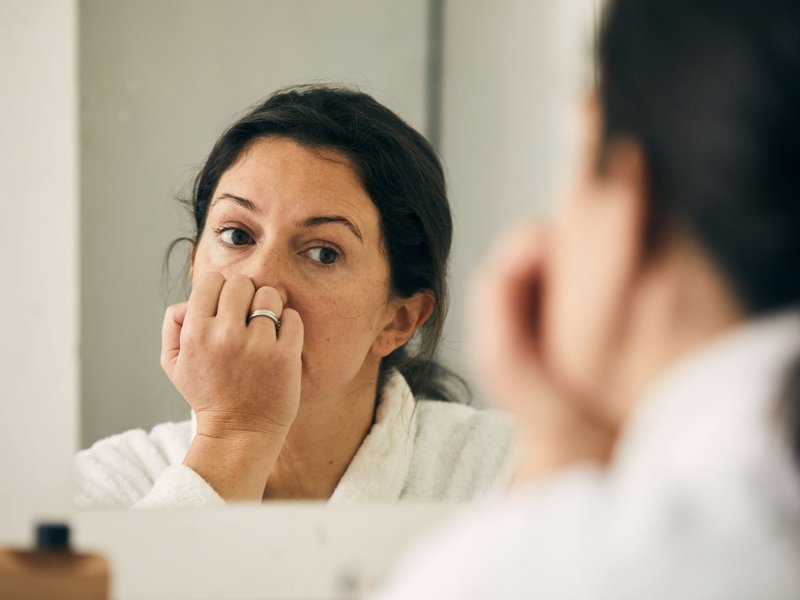A middle aged woman with dark hair stares anxiously into a mirror. There is a wedding band on her hand.