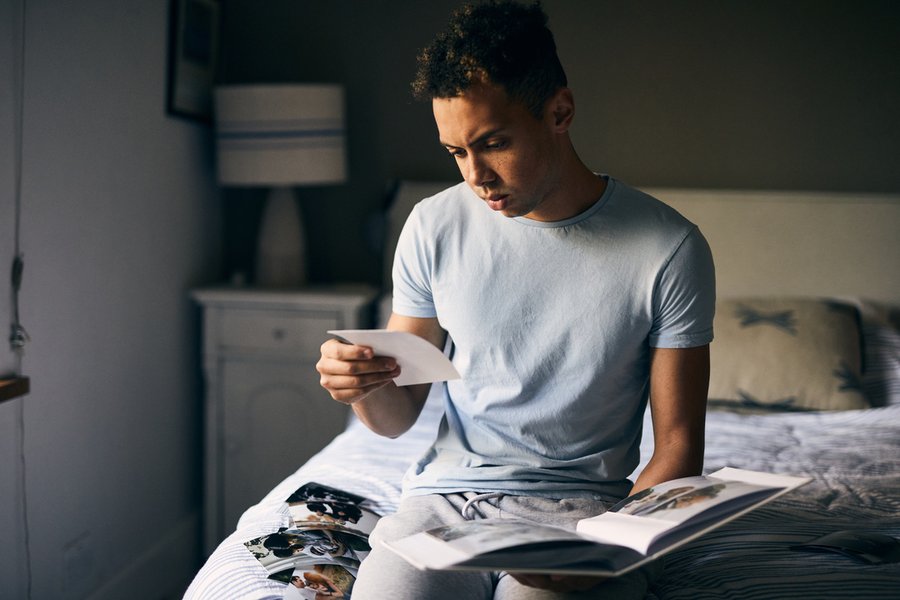 A young man sat on a bed is looking through a pile of photographs with a sad expression on his face.