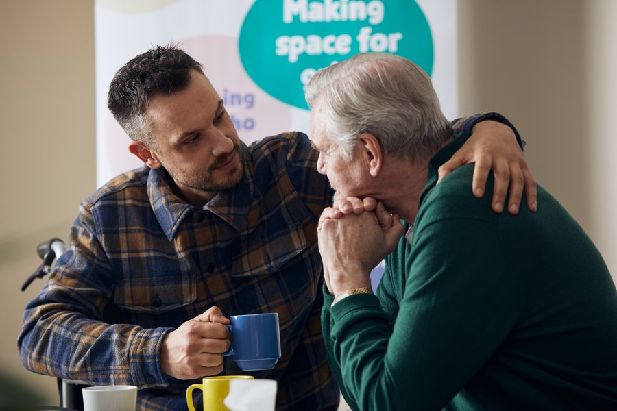 Two men are sat with mugs, the younger man has his arm around the older man in a comforting gesture