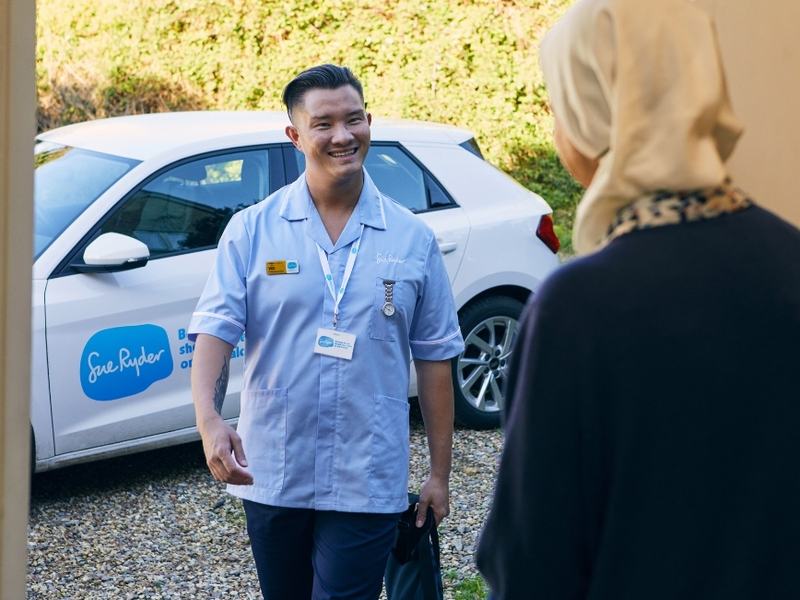 A Sue Ryder healthcare professional arrives at a person's home and smiles while greeting them at the front door, with a Sue Ryder care vehicle parked outside.