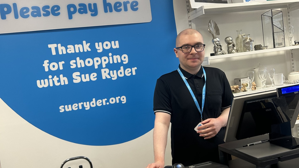 A person stands behind a shop counter. They are wearing glasses and have short hair. They smile gently towards the camera.