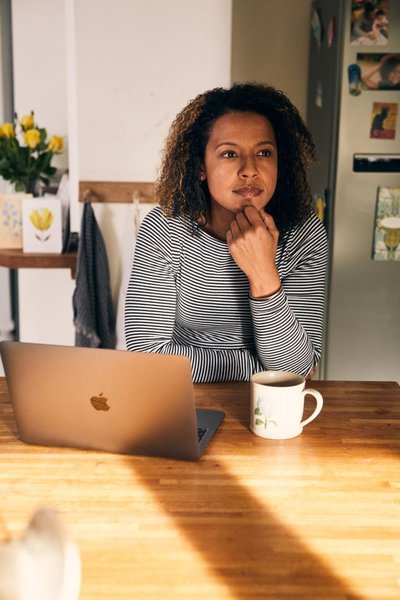 A woman is sat a table looking into the middle distance. In front of her on the table is a laptop and a mug.