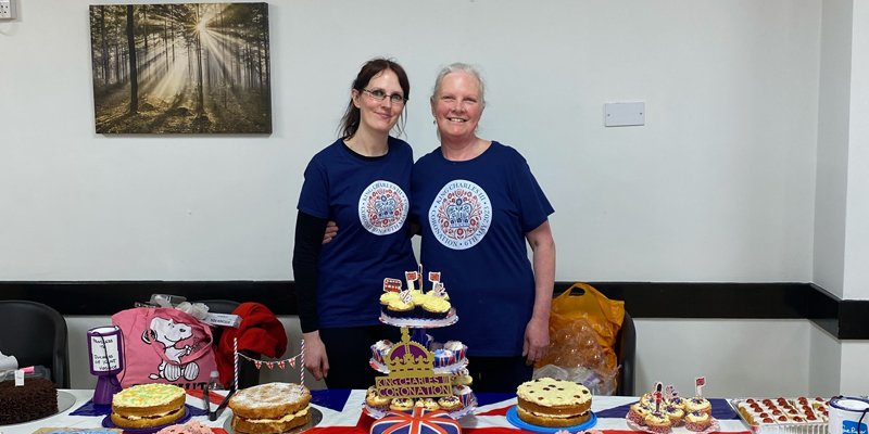 Two women standing behind a table covered in cakes, ready for their cake sale