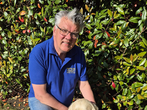A person with short grey hair and glasses kneels beside a golden retriever dog. They are outside on a grassed area, with foliage behind them.