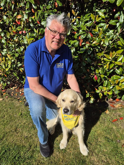 A person with short grey hair and glasses kneels beside a golden retriever dog. They are outside on a grassed area, with foliage behind them.