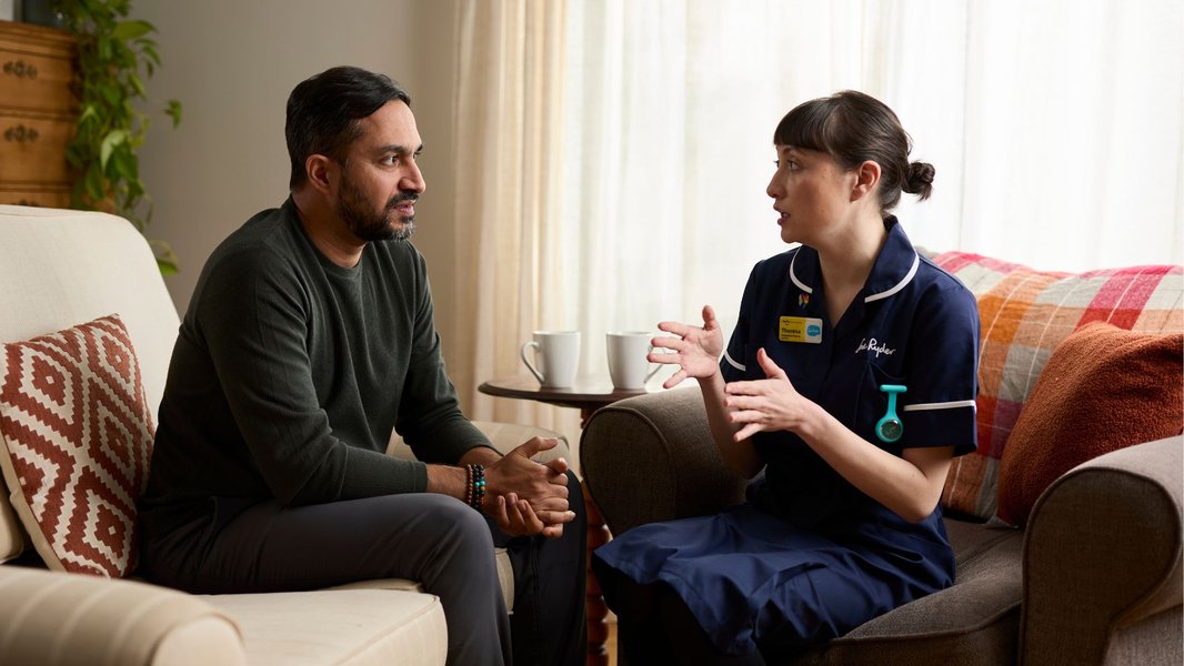 A man sits on a sofa listening to a Sue Ryder nurse, who is speaking and gesturing with her hands in a home setting