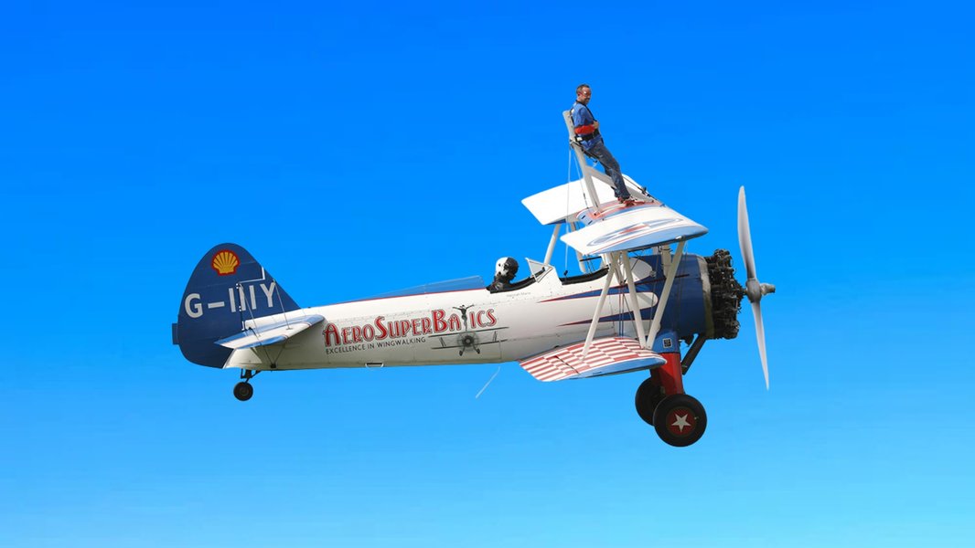 A person stands confidently on a biplane in flight, arms outstretched, with smoke trailing behind. The sky and a grassy field are in the background.