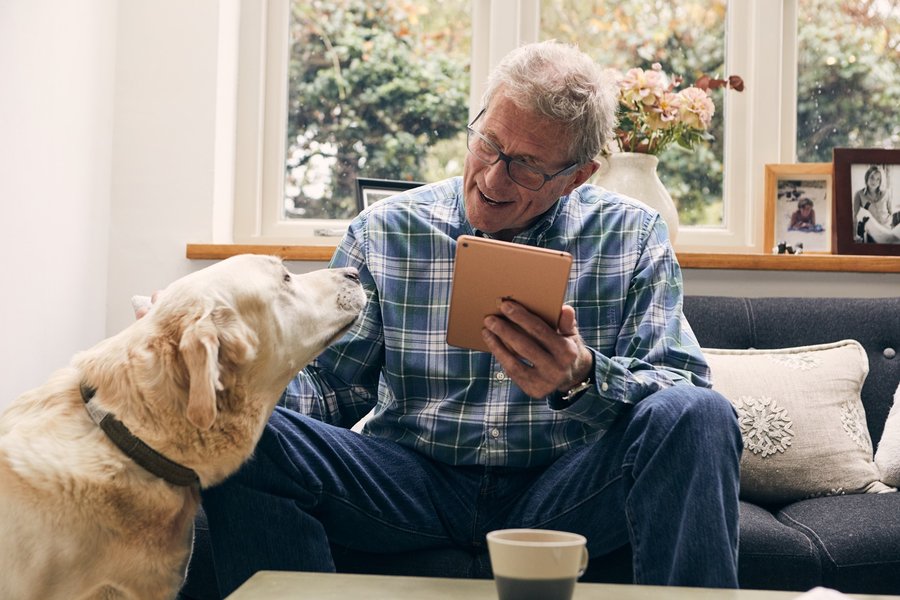 An older man is sat on a living room sofa holding an ipad and with a mug on the table in front of him. He's looking at a yellow dog at his side and has his hand up to pet the dog.