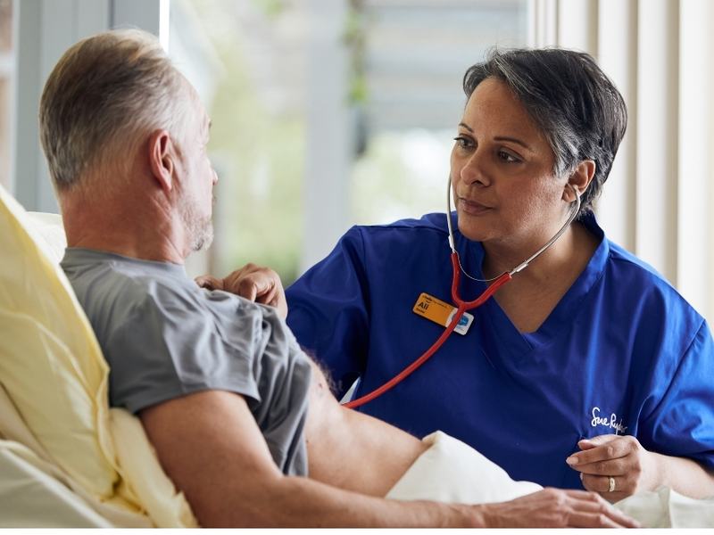 Sue Ryder nurse using a stethoscope to examine a patient in bed while providing hospice care.