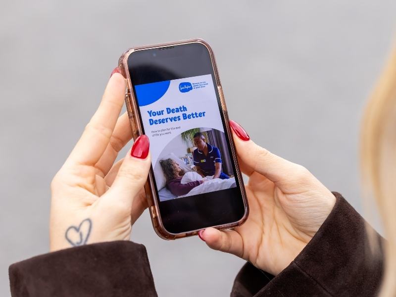 Person holding a smartphone displaying a Sue Ryder guide titled ‘Your Death Deserves Better’ about end-of-life care