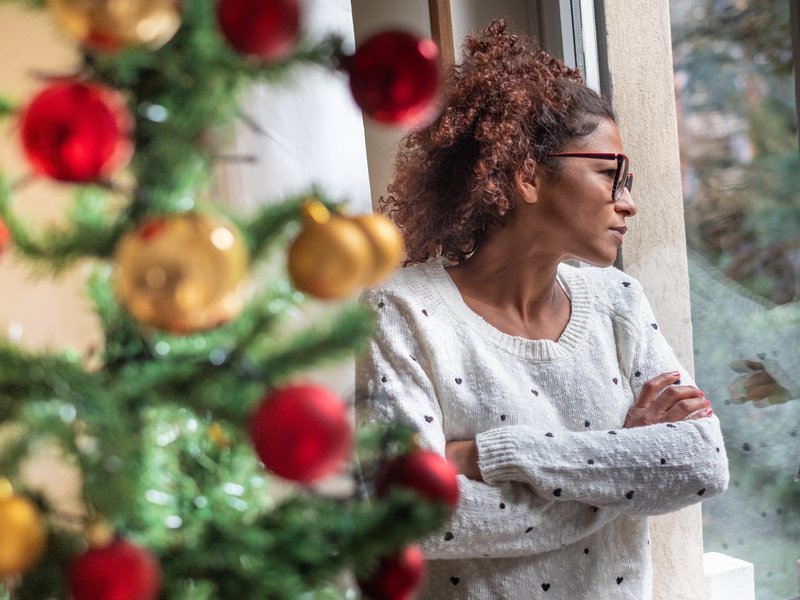 A black woman wearing glasses and a white jumper with spots is staring pensively out of the window with her arms folded.  In the foreground, there is a close up of a Christmas tree.