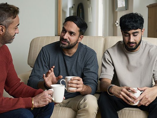 A dad and his two sons sitting in their living room with cups of tea, talking about a loved one they've lost