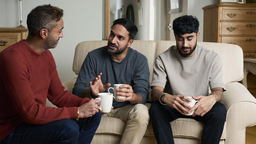 A dad and his two sons sitting in their living room with cups of tea, talking about a loved one they've lost