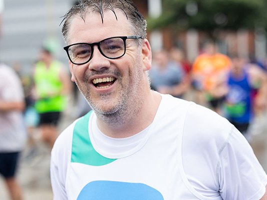 A Sue Ryder runner wearing glasses, smiling as he runs the race among other competitors