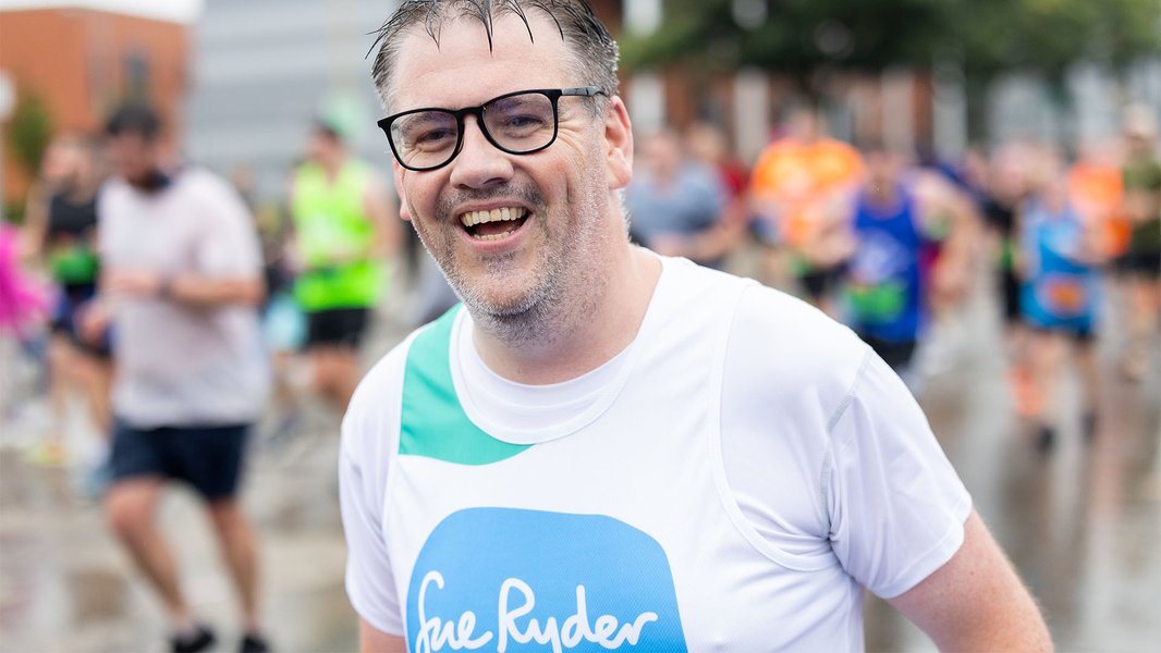 A Sue Ryder runner wearing glasses, smiling as he runs the race among other competitors