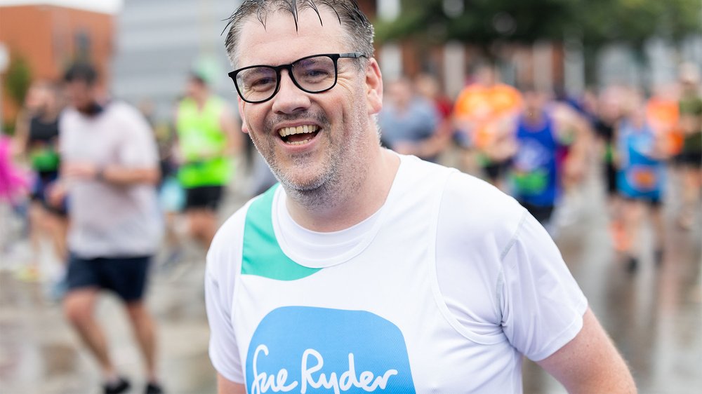A Sue Ryder runner wearing glasses, smiling as he runs the race among other competitors