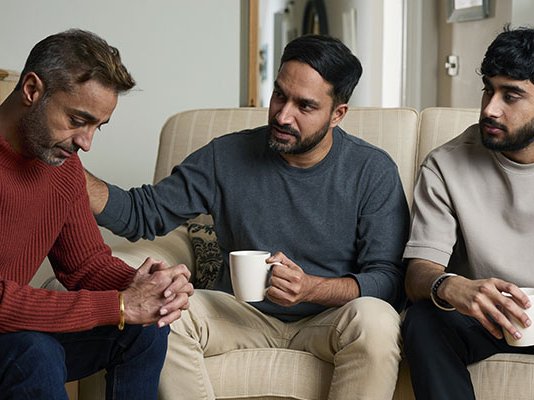 A grieving family together in their living room, with one of the sons comforting their father