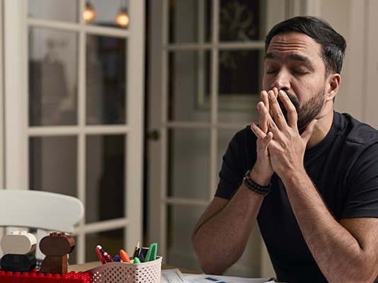 A grieving man with his eyes closed, holds his head in his hands while seated at his dining table alone