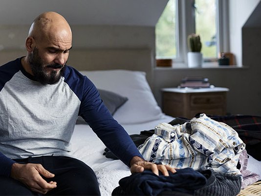A grieving man sitting on a bed, sorting through a loved one's clothing