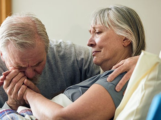 A patient lies in bed, as her husband cries at her bedside holding her hand