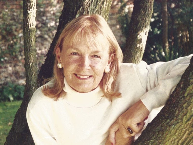 A woman with light hair, wearing a white turtleneck and pearl earrings, smiles while leaning against a tree outdoors.