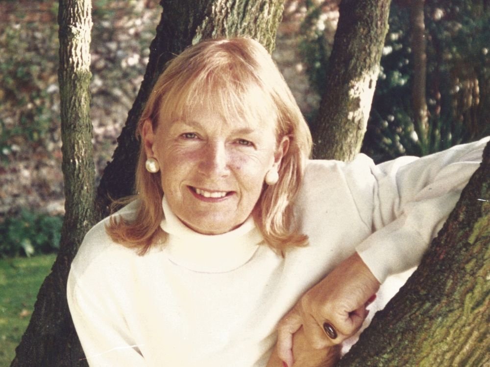 A woman with light hair, wearing a white turtleneck and pearl earrings, smiles while leaning against a tree outdoors.