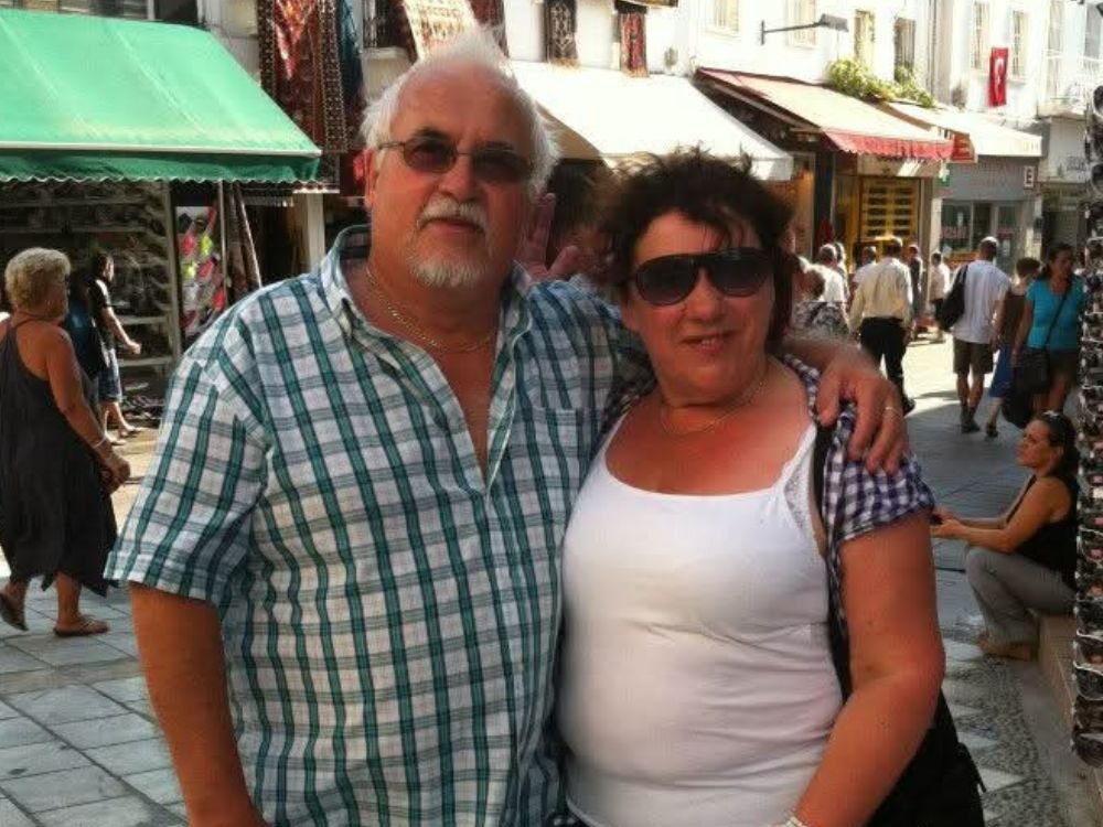 An older man and woman stand together on a street, smiling at the camera, with buildings visible in the background.