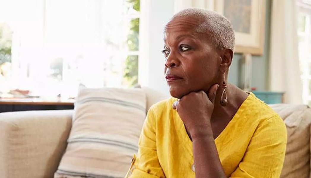 A black woman with cropped grey hair is sitting on a yellow sofa looking pensive.