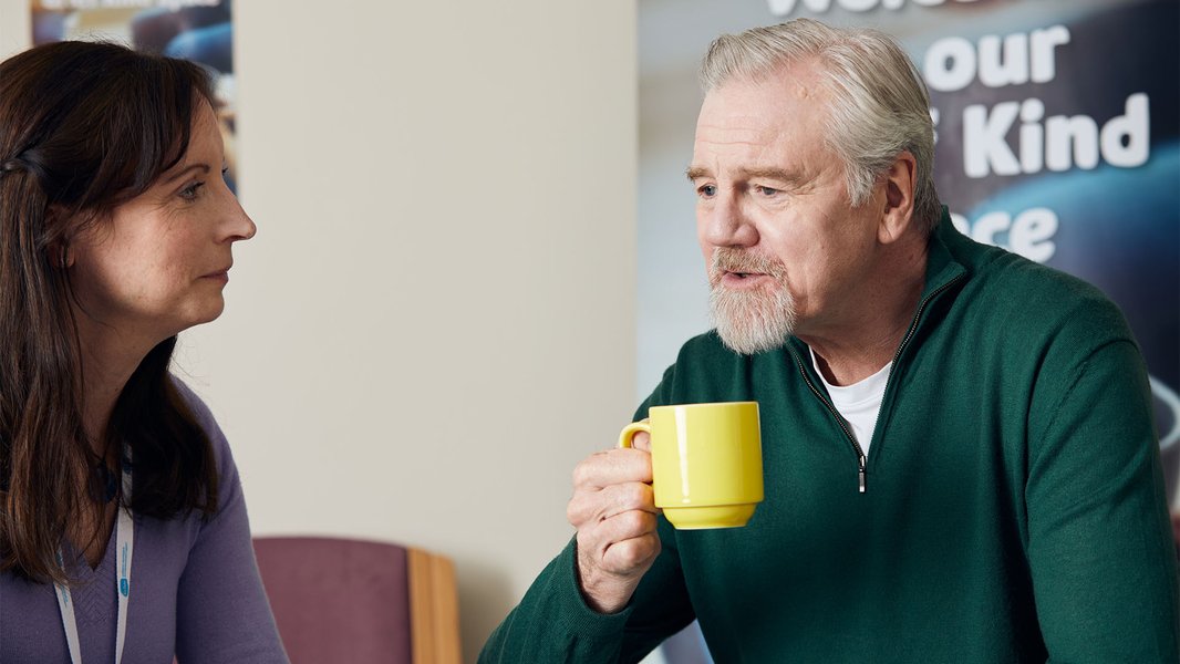 A volunteer talking with a man holding a cup of tea, at one our of Grief Kind Spaces