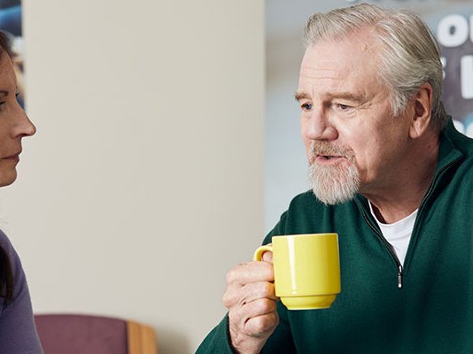A volunteer talking with a man holding a cup of tea, at one our of Grief Kind Spaces