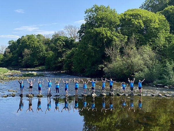 A line of walkers on stepping stones across a large river, with their reflections beneath them