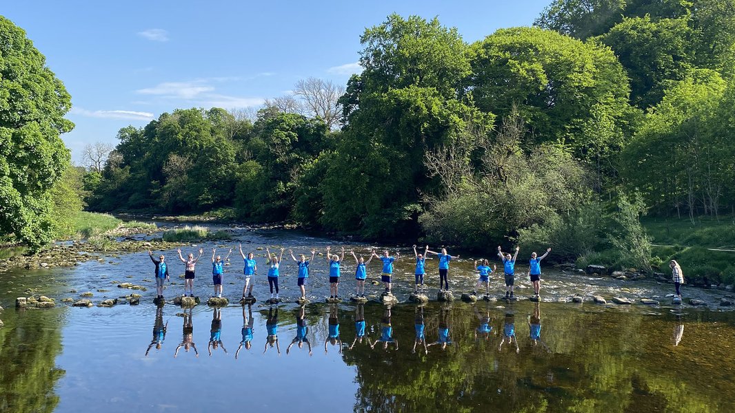 A line of walkers on stepping stones across a large river, with their reflections beneath them