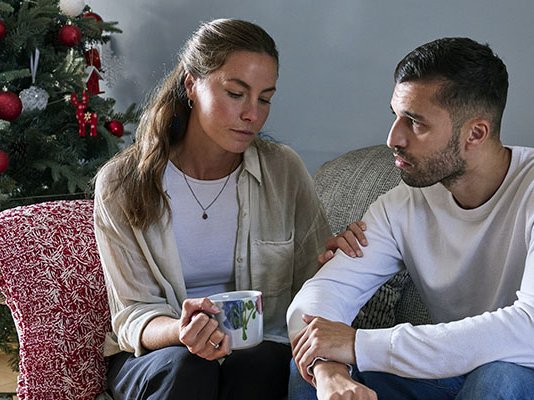 A woman consoling her grieving partner on the sofa, with a Christmas tree behind them
